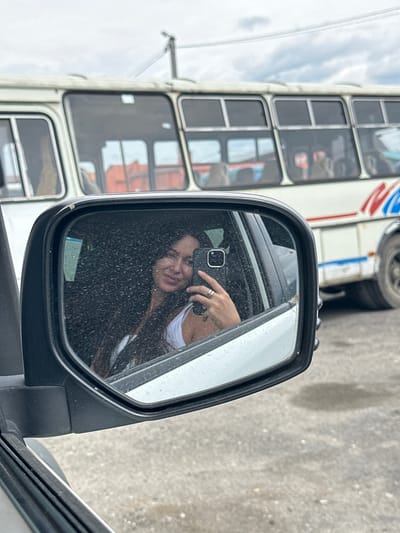 The image shows a woman taking a selfie in a car's side mirror. She is wearing a white tank top and has long, dark hair. She is holding a smartphone in her right hand and wearing rings on her fingers. In the reflection of the mirror, a white bus with red and blue stripes is visible in the background. The environment appears to be an outdoor setting, possibly a street or a parking lot, with a cloudy sky overhead. The specific location in the city of Slyudyanka, Russia, is not identifiable from the visual information provided in the image.