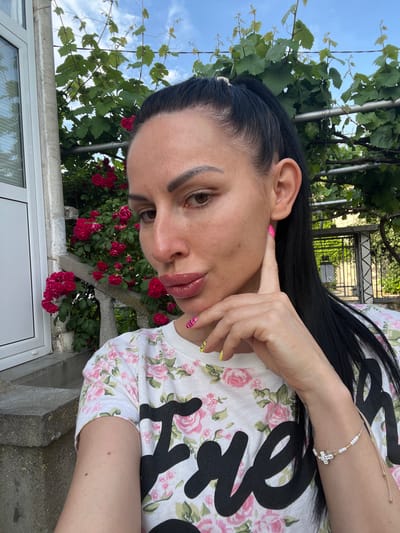 An adult female is captured in a selfie, positioned outdoors in Sungurlare, Bulgaria. She has long, dark hair styled in a ponytail, dark eyebrows, and wears pinkish-nude lip gloss. Her left hand is raised, with the index finger touching her cheek near her ear. Her fingernails are painted with a combination of yellow and black stripes and pink and black stripes. She is wearing a white t-shirt patterned with pink roses, green leaves, and partial black text reading "Fre" and "sh". On her right wrist, a beaded bracelet with white and gold beads and a sparkling silver-toned cross charm is visible.
The background reveals a verdant outdoor setting. To the left, a white window frame with interior blinds and a light-colored concrete structure are visible. Behind the subject, a dense bush with numerous red roses blooms next to a concrete balustrade or railing. Overhead, a metal trellis supports a grapevine with green leaves and tendrils. Further in the background to the right, a dark metal gate with an ornate pattern is partially visible through more foliage. The sky above is blue with scattered white clouds.