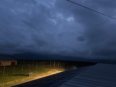 The image depicts a rural scene at dusk, likely in or near the city of Sisian, Armenia. In the foreground, the corrugated metal roof of a structure, possibly a barn or industrial building, is visible, covered in part by solar panels. A thin black wire stretches diagonally across the upper portion of the frame. Beyond the roof, a field with scattered white posts and a long, dark structure resembling a vineyard or orchard stretches across the mid-ground. To the left, a low, stone building with a wooden door and a metal roof is partially illuminated by a spotlight. In the background, dark, hazy mountains form a silhouette against the dramatic, cloudy sky. The overall lighting suggests either early morning or late evening, with artificial light from a spotlight illuminating a portion of the field and the vineyard. There are no people visible in the image. The environment appears to be agricultural and somewhat remote.