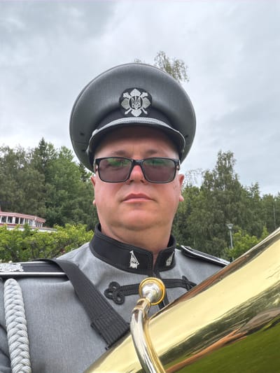 A man in a grey military-style uniform with a black collar and silver trim, a peaked cap with an emblem, and dark-rimmed glasses, is holding a large brass instrument, possibly a tuba. He is facing forward, with the instrument partially obscuring the right side of the frame. In the background, there are lush green trees, some foliage, and a building with a pink facade. The sky is overcast. The scene appears to be outdoors, possibly in a park or garden setting. The man's expression is neutral. The image does not contain any elements that specifically indicate the location is Aneby, Sweden.
