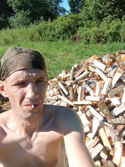 The media file is a selfie taken outdoors. The main subject is a man, seen from the chest up, wearing a green bandana and smoking a cigarette. His expression is neutral. Behind him, to the right, is a large pile of chopped firewood. The background consists of green grass and dense trees under a clear blue sky, suggesting a sunny day. The location is identified as Светласельскі сельскі Савет, Belarus. There is no visible text in the image.