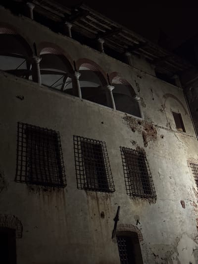The image shows the exterior of a building at night. The building has a rough, weathered facade with peeling plaster and exposed brickwork in places. There are several windows with heavy, dark metal bars. Above these barred windows, there is an arched colonnade with red brick arches supported by columns. The roof structure above the colonnade is visible and appears to be made of wood. The overall impression is one of age and disrepair. This building is located in Lagnasco, Italy.
