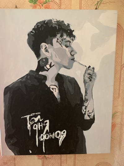 This is a painting of a person in a grayscale style. The subject is shown from the chest up, with their head tilted back and eyes closed. They are holding a cigarette or similar object to their mouth. The person is wearing a black shirt with white text on it. The background is a light gray color, with some abstract shapes in a lighter shade of gray. The overall mood of the painting is somber or introspective.

The visible text on the shirt appears to be in a stylized script. While the exact words are difficult to decipher due to the artistic rendering, it has a calligraphic quality.

The painting is placed on a surface that appears to be a table with a textured, patterned tablecloth featuring floral motifs. The tablecloth has a warm, peachy-pink tone with hints of green and brown. The setting does not provide any specific location cues beyond it being indoors. There are no indications of the time of day or weather conditions.