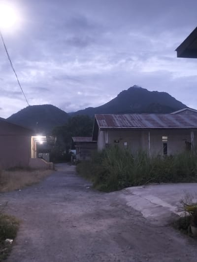 This media file captures an outdoor scene at dusk or dawn, with a mountain range looming in the background under a cloudy sky. The foreground features a dirt and gravel pathway winding between two buildings. To the left, a building with a light-colored wall is partially visible. To the right, a single-story building with a corrugated metal roof is prominent, showing several windows, some illuminated. Tall, green grass grows alongside this building and the pathway. The overall atmosphere is subdued, likely due to the low light conditions. There are no people or specific activities depicted, suggesting a quiet, possibly residential setting. The location cues, such as the architecture and vegetation, along with the visible mountain range, are consistent with a rural or semi-rural area in a tropical or subtropical region. The time of day is indicated by the diffused light and the cloud cover.