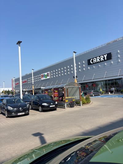 The image depicts a sunny outdoor scene in front of a modern shopping center located in Modlniczka, Poland. The shopping center's facade is a light gray, paneled material with a repeating pattern of square indentations. Prominently displayed on the building is a large black sign with white lettering spelling out "CARRY." To the left of the "CARRY" sign, the building is also branded with "Smyk" in a colorful logo, and further to the left, "martes" is visible in red lettering.
In the foreground, several cars are parked on a paved lot. These include a black Renault Clio with license plate "KWI 5454F" and a black Volkswagen Fox with license plate "LC 84952." A green vehicle's hood and windshield are visible in the lower right and bottom of the frame, reflecting the shopping center and its surroundings.
Adjacent to the shopping center entrance, there is a covered area with shopping carts. Various advertisements are displayed on the sides of this structure, including one that appears to be for a food or butcher shop, featuring images of meat.
Several people are visible near the entrance of the shopping center. One person, wearing a yellow vest and a hat, stands near the entrance, seemingly directing or assisting customers. Other individuals are walking in and around the shopping center.
The sky is a clear, bright blue, indicating fair weather. Tall, white light poles are strategically placed throughout the parking area. The overall environment suggests a commercial district on a typical day.