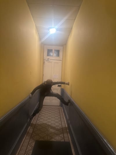 The scene depicts an interior hallway within an unidentified building in Roanne, France. The hallway features pale yellow walls above a dark blue-grey dado rail, with a geometrically patterned tiled floor. A bright, centrally mounted overhead light fixture provides illumination. At the end of the hallway, a white wooden door with two textured glass panels and a small affixed notice is visible.

A single individual is observed horizontally suspended across the hallway's width, with their back to the viewer. They are dressed in dark attire, including a long-sleeved top, pants, boots, and a patterned beanie. Their limbs are extended and braced: the left hand and foot against the left dado rail, and the right hand and foot against the right wall and dado rail respectively, maintaining an elevated position between the opposing surfaces.