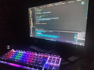 A dark indoor scene is depicted in Rozovo, Bulgaria, featuring a computer workstation. A widescreen computer monitor occupies the upper right portion, displaying programming code within an integrated development environment (IDE) against a dark background. Visible elements on the screen include a file explorer, syntax-highlighted code with references to configuration files and `app.module.ts`, and a terminal window at the bottom displaying command-line output. The taskbar in the lower right of the screen indicates application icons, including Spotify, and the time "04:00 AM". Directly in front of the monitor, a full-size mechanical keyboard illuminates the dark environment with active multi-colored RGB backlighting, showcasing a gradient of red, orange, yellow, green, blue, and purple across its keys. A dark, cylindrical object is partially visible on the left side, behind the keyboard, and another dark, indistinct object, likely a computer mouse, is partially visible to the right of the keyboard. No individuals are present in the image. The scene is characterized by low ambient light, with the monitor and keyboard backlighting serving as the primary light sources.