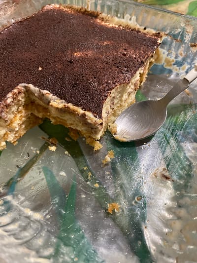 A partially eaten tiramisu sits in a clear glass dish. A silver spoon rests on the remaining portion of the dessert. Crumbs and residue from the tiramisu are scattered around the dish. The location appears to be indoors. There are faint green and blue hues in the background. There is no indication in the image that the location is Capesterre-Belle-Eau, France.