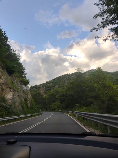 The image depicts a winding asphalt road viewed from inside a vehicle, likely a car, heading towards a mountain pass. The road is bordered by metal guardrails, and rocky cliffs with vegetation on the left, and forested hills on the right. The sky is filled with partially cloudy weather. The location is plausibly near Gospodintsi, Bulgaria, given the terrain and general road infrastructure. No people are visible.