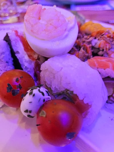 This image captures a close-up of an individual's plate of assorted appetizers within an indoor dining establishment in Audun-le-Tiche, France. The scene is illuminated by a distinct purple ambient light. Dominating the center-top is a hard-boiled egg half, white in color, topped with a single pink, cooked shrimp. To its immediate left are partially visible sections of sushi rolls featuring white rice and dark nori. To the right of the egg, a mound of finely chopped, reddish-brown food, possibly a tuna or meat salad, is visible, partially topped with an orange sauce. Below the egg and to the left, two red cherry tomatoes are present, one exhibiting minor dark spots. Between these tomatoes and a large, oblong rice-covered item (likely another sushi roll or rice ball) is a small, white, ovoid object speckled with green herbs, resembling a mozzarella ball. A piece of salmon sushi is partially visible towards the upper right. The food is resting on a white plate. The background is out of focus, showing blurred lights and shapes characteristic of a restaurant or buffet setting. No individuals are visible.