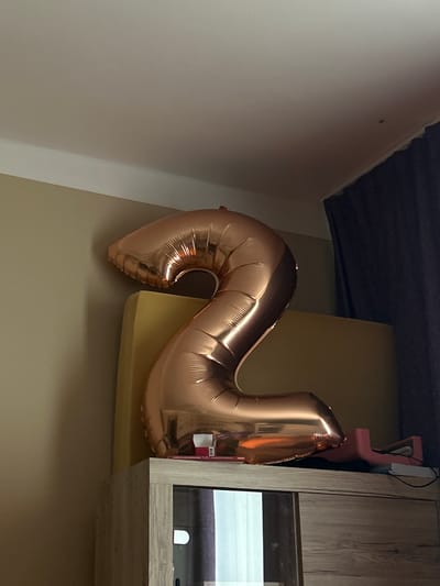 This media file captures a close-up view of a rose gold number "2" balloon, inflated and casting a slight shadow. The balloon is the primary subject, positioned in the foreground and dominating the frame. It rests on what appears to be a yellow cushioned surface, possibly a sofa or chair.
In the background, a light-colored wall forms the left boundary of the image, transitioning to a darker, angled surface at the top, which could be a ceiling or an overhang. To the right, a wooden cabinet with a glass-fronted compartment is visible. Within the cabinet, a white, arch-shaped light is illuminated. The overall lighting suggests an indoor setting, likely during daylight hours, though artificial light from the cabinet is also present.
A pink object, possibly another decorative item or part of a toy, is partially visible behind the balloon and next to the cabinet. There is a small piece of white paper attached to the balloon with some red text and a barcode, though the text is not legible. No people are visible in the image. The scene is static, with no discernable activity taking place. The composition, with the balloon prominently featured and the background elements providing context, suggests a celebratory or decorative arrangement. The location is Teplice, Czechia.