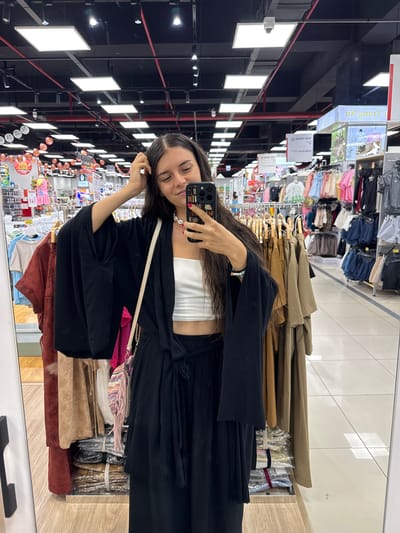 A young woman with long, dark hair is taking a mirror selfie in what appears to be a clothing store. She is wearing a white crop top and a black kimono-style top with wide sleeves, tied at the waist with a black sash. She also has on black pants. A patterned bag with fringes is slung across her body. She is looking at her phone, which she is holding in her right hand, and using her left hand to lightly touch her hair. The background shows racks of clothing, display shelves, and overhead lighting. The overall impression is that of a shopping trip. The image is in focus and well-lit, with no discernible location in Phu Thuy, Vietnam.