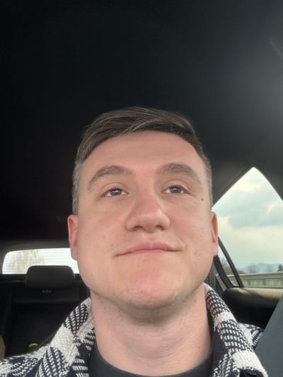 The image captures a close-up, selfie-style view of a male individual seated within a vehicle. The subject is a light-skinned adult with short brown hair, wearing a black and white patterned outer garment over a dark undershirt. His eyes are directed upwards and slightly to his left, with a neutral to slight smiling expression. The vehicle's dark headliner is visible above him. Through the window to his right, a cloudy daytime sky is visible above distant hills or mountains, with a grey barrier or guardrail present below. To his left, a black headrest is visible in the background, partially obscuring the view through a rear window. No specific landmarks are present in the image to confirm the location as Aichdorf, Austria.