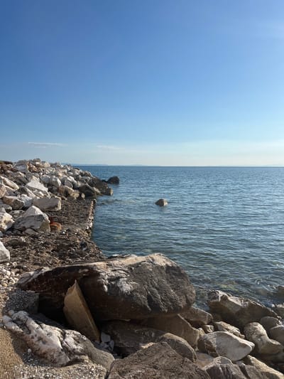 The image depicts a rocky shoreline on a clear day. The foreground is dominated by large, irregularly shaped rocks, some of which are white with dark veining, and others are dark grey. A path or rough area made of smaller stones and dirt leads towards the water. On the left side of the path, a pile of large white rocks is visible, some appearing to be artificial additions to the natural landscape. A rusty metal object is partially submerged in the ground near the water's edge.
The water is a calm, clear blue with gentle ripples. A few scattered rocks are visible in the water, with one larger, darker rock positioned to the left of center. In the distance, a hazy line of land is visible on the horizon, suggesting other islands or the mainland. The sky is a bright, cloudless blue, fading to a lighter shade towards the horizon. The overall impression is of a natural, somewhat rugged coastal environment. The location is described as the city of Potos, Greece.