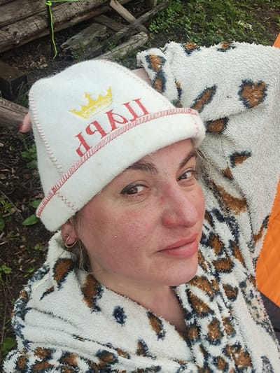 A middle-aged woman is seen lying down outdoors in Полянки, Russia. She is wearing a white terrycloth sauna hat with a red inscription, partially obscured, but featuring a yellow crown. The hat's trim is stitched in red. She is wrapped in a white and brown patterned fleece blanket. Her head is turned towards the camera. The background includes greenery, possibly grass and foliage, and fragments of what appear to be rough-hewn wooden structures or logs. Part of an orange material is visible in the upper portion of the image.