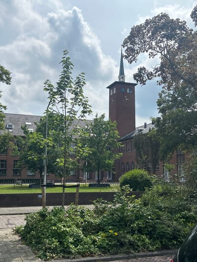 The image shows a brick building with a prominent clock tower topped with a green spire. The building has a red brick facade, a grey tiled roof with several dormer windows, and numerous tall, arched windows. The clock tower has round clock faces on at least two sides. Lush green trees and bushes are visible in the foreground and around the building. The sky is partly cloudy. A street and sidewalk are in the lower portion of the image. The location is the city of Leiden, Netherlands.