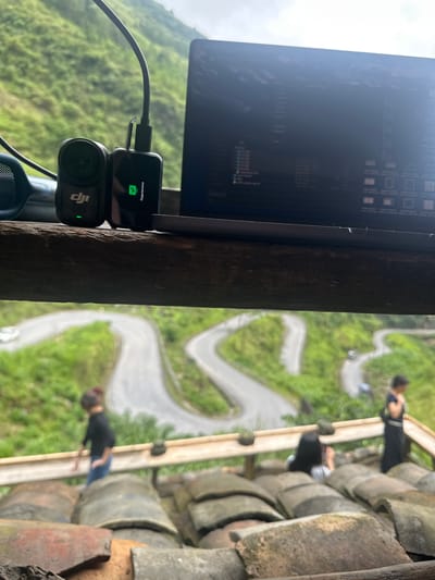 The image shows a laptop with a DJI action camera connected, positioned on what appears to be a wooden beam or ledge. The laptop screen displays program code or file directories. Through the background, a winding road through lush green terrain is visible, suggesting a mountainous or hilly location. On the left side, a roof made of tiles is seen, and several blurred figures can be distinguished walking on the roof with a winding road behind them. Based on the provided information, this scene is located in Yên Minh Commune, Vietnam.