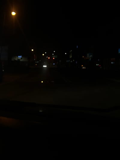 The image captures a blurry nighttime scene, likely from inside a vehicle looking out at a city street. A prominent street light with a yellowish-orange glow is visible in the upper left quadrant. In the distance, there are out-of-focus lights from buildings, traffic lights, and possibly other vehicles. The overall impression is one of low visibility and ambient light from artificial sources against a dark backdrop. The environment appears to be a street in Siedlce, Poland, characterized by typical urban street lighting and building structures. No individuals or specific actions are discernible due to the blurriness and low light conditions.