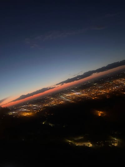 The image displays an aerial or high-vantage-point view of the city of Beni Mellal, Morocco, at dusk or early night. The horizon is tilted upwards to the right, creating a prominent diagonal composition.
The upper portion of the scene features a dark navy blue sky transitioning to a lighter blue-grey, with a distinct, elongated band of vibrant orange-red light along the horizon, indicative of a sunset or twilight glow. Scattered, dark, elongated cloud formations are present, primarily above and slightly within this orange-red band.
The middle ground is occupied by the extensive sprawl of Beni Mellal, illuminated by numerous warm-toned, bright city lights. These lights appear as diffuse, yellow-orange specks, suggesting streetlights and building illumination, extending across the landscape into the distance.
The foreground, occupying the lower third of the image, is predominantly dark and appears to be the rugged, unlit terrain of a mountain or hill immediately below the viewpoint. A few isolated, faint lights are discernible within this dark foreground, indicating sparse structures or distant pathways. No people, specific subjects, or actions are visible within the frame. The overall visibility of the distant cityscape lights is clear, despite some atmospheric diffusion.