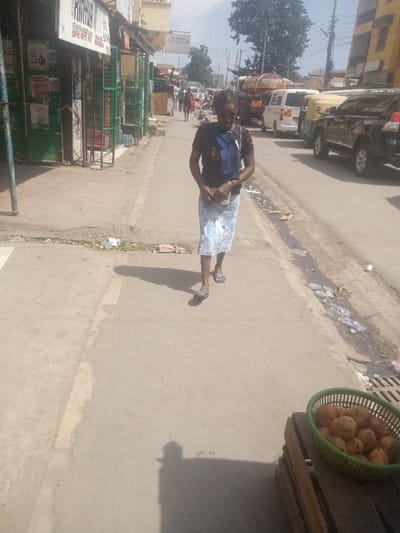 A woman walks on a sunlit sidewalk in an urban area. She carries a blue bag over her shoulder and wears sandals. Trash lines the side of the road. A green plastic basket filled with brown fruit sits on a wooden pallet in the foreground. Cars and other people are visible in the background. Stores line the street.