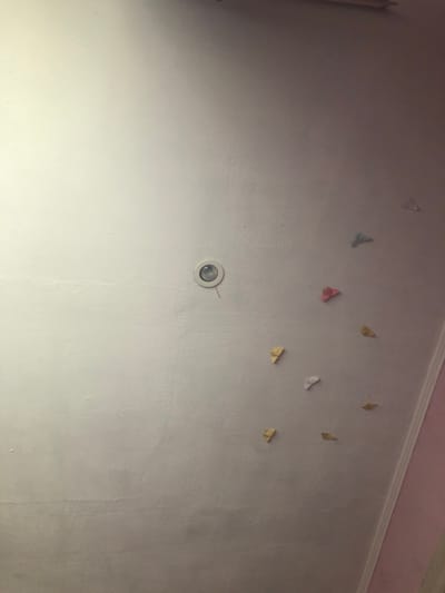 The image shows a white wall with several small, colorful paper butterflies adhered to it. In the upper right quadrant of the image, a white recessed ceiling light is visible. The lighting in the room appears to be coming from the ceiling light and possibly an unseen window, casting a shadow on the right side of the wall. The wall's texture is slightly uneven, with some visible imperfections. The overall scene suggests a decorative element in a room, possibly a child's bedroom. No people or specific locations in Baryshivka, Ukraine are discernible.