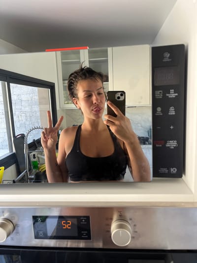 A female individual is depicted in a selfie-style reflection on the polished door of a built-in kitchen microwave oven. She is centrally positioned, holding a dark-colored smartphone in her right hand, pointed towards herself. Her left hand is raised in a "peace" sign, and her tongue is slightly extended from her mouth. She has short, dark hair and wears a black sports bra.
The reflected background reveals a modern kitchen interior, featuring white upper cabinets and a light-colored countertop. A large window with a black frame shows an exterior wall constructed from grey, irregular stone blocks. A kitchen sink area with a flexible pull-out faucet and a green liquid dispenser is visible to the left of the window.
To the right of the reflection, the microwave's black control panel displays "00:00" and several touch controls labeled in Spanish, including "Smart Inverter." Directly beneath the microwave, a stainless steel conventional oven is visible, its digital display showing "52," flanked by rotary control knobs. This scene is located within a residence in La Barra, Uruguay.
