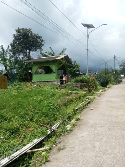 This image depicts a semi-rural outdoor scene in Ura-Dozhi, Bhutan, under an overcast sky. A prominent lime-green, open-sided wooden or bamboo shelter with a corrugated metal roof is centrally positioned on a raised, overgrown area. To its left, a tall, dark-leaved tree stands amidst lush green vegetation, including banana plants. Adjacent to the shelter's right, a tall metal pole supports a solar panel and a streetlight fixture. Multiple overhead power lines crisscross the sky.
Two individuals are visible near the green shelter; one, wearing a light top and dark bottom, stands with their back to the camera, partially obscured by the shelter's right side, seemingly interacting with an object or space. The second individual is mostly hidden behind the first. To the left of the shelter, a rectangular yellow utility box or container is partially visible.
In the foreground, a concrete path or narrow road extends from the lower right towards the distant background, flanked on its left by an overgrown ditch or embankment. White PVC pipes are laid horizontally along this embankment, some supported by wooden planks, partially obscured by dense green foliage. Further along the concrete path, a small yellow item lies on the ground. Stone steps and a retaining wall separate the elevated area of the shelter from the lower ground level. Distant hills or mountains are visible under the cloudy sky, and additional utility poles with wires are present further down the road.