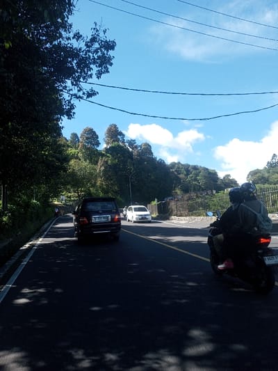 This daytime image depicts a paved two-lane road located in Ka Thaung (Upper), Myanmar, traversing a densely vegetated, hilly landscape under a clear blue sky with scattered white clouds. The road surface is dark asphalt, marked with a white solid line on the left shoulder and a yellow solid center line. Tree shadows are cast upon the road. In the immediate foreground on the right, two individuals are riding a black motorcycle, both wearing helmets; the passenger wears a grey backpack and pink shoes. The motorcycle displays the Indonesian license plate B 6629 VXA. Ahead on the left, a dark SUV with the Indonesian license plate B 3344 KXA is visible, followed by a white sedan. Dense tropical foliage and large trees line both sides of the road, with a steep, tree-covered hillside dominating the background. Power lines run horizontally across the upper portion of the frame. A stone wall and black metal fence are visible on the right side of the road in the mid-distance.