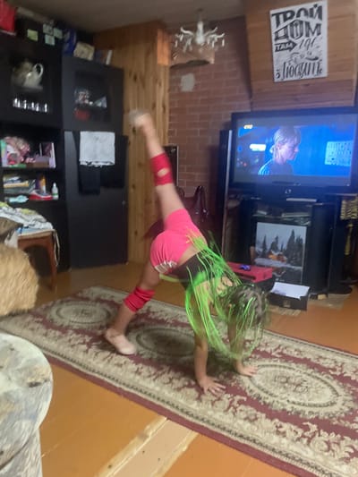 The image depicts a young person with long, bright green hair in a split, stretching on a patterned rug. They are wearing pink shorts with white trim and red knee pads. The setting appears to be a living room with a television playing a program that shows a person with blond hair. A brick wall is visible behind the television, and there are shelves with various items, including books and what looks like a tea set. The overall lighting suggests it's daytime, and the room is a bit cluttered, suggesting a home environment. There is text on a sign on the wall above the television that reads "ТРАЙ ДОМ" and "Я ЛЮБИТЕ". The scene is captured from a slightly elevated, oblique angle.