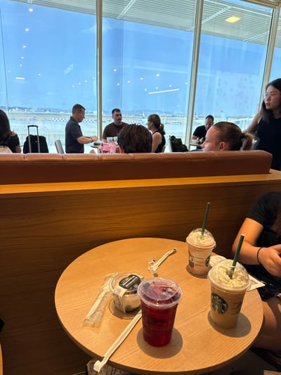 In a brightly lit coffee shop with large windows overlooking an airport tarmac, a group of people are gathered around a circular wooden table. The scene is set during the daytime, likely morning or afternoon, with ample natural light streaming in. The atmosphere appears relaxed, with individuals engaged in conversation or enjoying their beverages.
Dominating the table are three drinks: two iced coffee beverages with whipped cream and straws in clear plastic cups bearing the Starbucks logo, and a larger cup filled with a red, berry-like drink, also with a straw. Beside the red drink, a small, clear plastic container with a black lid sits on a napkin. A white paper straw in a plastic wrapper lies next to it.
The people in the foreground are mostly out of focus, but several individuals are visible. A woman with her hair in a ponytail is looking down, and another person's arm with a bracelet is resting on the table. In the background, through the expansive windows, an airport scene unfolds. Airplanes are visible on the tarmac, and a man in a dark shirt is looking out at the view. The sky is a clear blue. The coffee shop appears to be an airport lounge or café, indicated by the presence of luggage, including a black rolling suitcase, and the airport environment visible through the windows. The wooden interior of the coffee shop, with its booth seating and table, provides a warm and inviting setting.