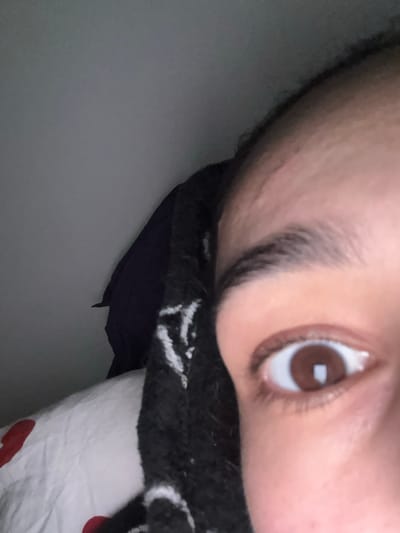 A close-up image shows a person's face tilted at an angle. The right eye is in focus with a brown iris and a white highlight reflecting light. An eyebrow sits above the eye. Part of a black and white patterned scarf or garment is visible below the face. Behind the scarf, a dark blue or black garment is visible. The background is a light gray, potentially a wall.