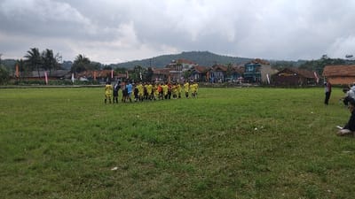 This wide-angle outdoor photograph captures a community event, likely a soccer match, on a large green grass field in Kecamatan Panjalu, Indonesia. In the middle ground, two groups of individuals, presumably soccer teams, are gathered. One team wears bright yellow jerseys with dark shorts, while the other is dressed in dark-colored (possibly black or dark blue) jerseys. They appear to be interacting, possibly engaging in a pre-match handshake or discussion, with a few individuals who might be referees or coaches among them.

To the right, closer to the foreground, a smaller group of people, likely spectators or officials, are visible, some standing and others crouching or sitting on the side of the field. The background showcases a rural or semi-rural village scene with numerous houses and buildings of varying styles and colors. Prominently featured is a brown building with white lettering on its facade, partially readable as "BALAI DESA," which translates to Village Hall, indicating the local government or community center. Beyond the village structures, a range of rolling hills or mountains stretches across the horizon. The sky is overcast with thick clouds, suggesting a cloudy day rather than bright sunshine. The field itself is an open, grassy expanse, typical of a local sports ground. The image portrays a local sporting event taking place within a distinct village and natural landscape.