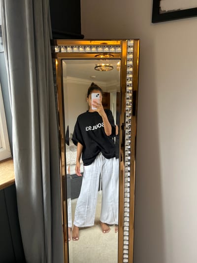 The image displays an interior scene located in Rushden, United Kingdom, featuring a female individual captured in a full-length, ornate mirror while taking a selfie. The person, with her hair styled in a bun and appearing barefoot, is wearing an oversized black t-shirt with the reversed white text "səɔuoe" and loose-fitting white wide-leg pants with a visible white drawstring. She holds a light-colored smartphone, which partially obscures her face, with a pink-toned case. A ring is visible on her right ring finger. The mirror itself features a prominent gold-colored frame adorned with numerous small, square, reflective or faceted inserts. To the left of the mirror, a window with a white frame and wooden sill is partially visible, covered by heavy light-grey curtains, with a dark panel or furniture item below the sill. To the right, a plain light-grey wall is visible, with a black-framed object positioned higher up. The reflection in the mirror reveals a room with light-colored carpeting, a ceiling featuring crown molding, and a circular ceiling light fixture (possibly a chandelier). Additionally, glimpses of a bed or sofa with light-colored upholstery and a dark piece of furniture are visible in the reflection.