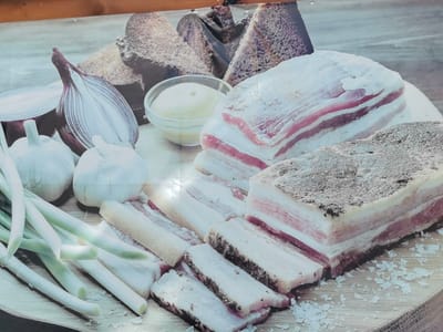 The image displays a still life arrangement of food items, possibly a depiction of local Pinsk cuisine. The foreground features a bundle of green onions, two heads of garlic, and a halved red onion. Behind these, several cuts of cured pork fat (salo) are prominently displayed, some whole and some sliced, revealing distinct layers of fat and meat. A small glass bowl containing a pale yellow condiment, possibly mustard or a similar spread, is visible. In the background, slices of dark bread are stacked, suggesting a rustic meal setting. The items are arranged on a wooden surface, with scattered white crystals, likely salt, on the board. The overall lighting suggests an indoor setting, possibly a food market or a display. There are no people or actions depicted in the image.