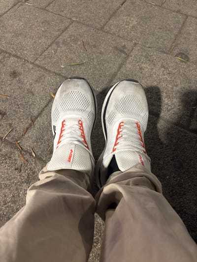 The image shows a pair of white athletic shoes with orange laces, worn by an unseen person standing on a paved surface. The shoes appear to be made of mesh material and have the brand name "ACRIVIT" visible on the heel tab of the left shoe. The paving stones are gray and weathered, with a few fallen leaves scattered on the ground. The lighting suggests it is nighttime or dusk, casting shadows on the ground. The image does not provide any specific information about the location in the city of Toulon, France.