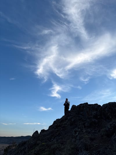 The image shows a low-angle shot of a man standing on a rocky precipice. The sky behind him is blue with wispy white clouds. Below the rock formation is a view of rolling hills and a city in the distance. Based on the location in Невон, Russia, it can be assumed this is a natural setting. The man is dressed in dark clothing and appears to be holding something in his hands.