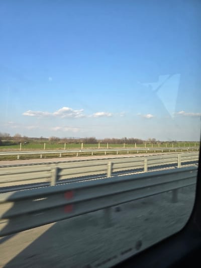 This image depicts a highway scene within the vicinity of Opalchenets, Bulgaria. The perspective is from inside a moving vehicle, looking out through a side window.
The foreground displays a multi-lane asphalt or concrete highway surface with multiple parallel metal guardrails (barriers). The closest guardrail, positioned in the lower mid-ground, is silver-gray, consisting of horizontal beams supported by vertical posts. Two distinct red circular markings are visible on one of the horizontal beams of this guardrail. Further back, additional guardrails separate multiple lanes or shoulders. Beyond the highway infrastructure, the mid-ground reveals expansive green and brown fields, transitioning to a line of sparse, leafless trees or bushes along the horizon.
The upper portion of the image is dominated by a clear, bright blue sky with a few scattered white cumulus clouds hovering low on the horizon. A distinct, angular reflection is present on the upper right window surface, resembling a partial human figure or an interior object from the vehicle. No individuals are directly visible outside the vehicle. The scene is illuminated by natural daylight, casting shadows from the guardrails onto the highway surface.