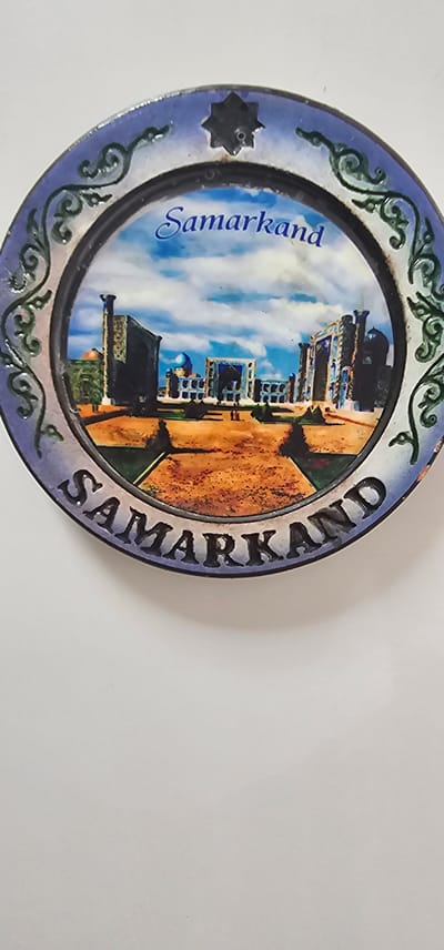 A circular decorative plate features a depiction of the Registan square in Samarkand, Uzbekistan. The plate has a blue and purple ornate border with a star motif. The central image shows several iconic ancient buildings with blue domes, tall minarets, and intricate tilework, set against a backdrop of a bright blue sky with white clouds. The ground is depicted as a reddish-brown dusty area. The name "Samarkand" is written in a blue cursive font across the top of the image, and the same name is embossed in capital letters around the bottom edge of the plate. There are no people visible in the image. The plate is presented against a plain white background.