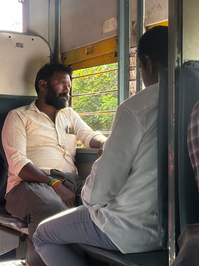 The image depicts two men seated inside a train compartment in Madirimangalam, India. The man on the left, with a beard and dark hair, wears a light-colored, patterned shirt and dark pants. He is looking out the window, which reveals a sunlit view of green foliage. His left arm rests on his leg, and he wears colorful bracelets on his wrist. A pen is tucked into his shirt pocket, alongside a mobile phone. The man on the right is seen from behind, wearing a white shirt. He appears to be looking in the same direction as the man on the left. The train interior is visible, with the yellow window frame and parts of the seating and partition. The lighting suggests it is daytime, with sunlight streaming in through the window.
