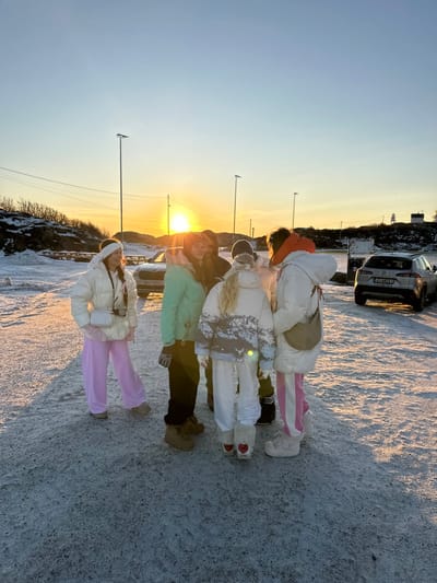 Six individuals are positioned on a snow-covered, unpaved surface in Henningsvær, Norway, during either sunrise or sunset, indicated by a low sun casting an orange glow across the horizon. The sky transitions from orange-yellow at the horizon to pale blue overhead.
All individuals are dressed in winter clothing. From left to right:
1. A person in a white puffer jacket, pink wide-leg pants, a white headwrap, and light-colored boots. A small white crossbody bag is worn, and a camera hangs from the neck. This individual faces slightly towards the viewer and smiles.
2. A person in a mint green puffer jacket, dark pants, and brown boots.
3. A person in a dark jacket, partially obscured by other individuals.
4. A person in a white puffer jacket featuring a grey and light blue mountain pattern, white pants, and white furry boots adorned with red heart emblems on the heels.
5. A person in a white puffer jacket with an orange collar/hood lining, pink and white striped pants, and light-colored boots, carrying a light brown crescent-shaped shoulder bag.
6. A person mostly obscured behind the individuals in the center of the group.
The individuals are clustered, indicating interaction. In the background, a light-colored SUV is parked behind the group, displaying a partially visible license plate beginning with "DH 4". To the far right, a silver SUV is parked with the Norwegian license plate "KJ 84215". Several tall utility poles with horizontal fixtures and power lines span the mid-ground. The landscape consists of rocky, snow-dusted hills with sparse, low-lying vegetation. A white lighthouse is visible on a distant hill to the far right. A body of water is visible beyond the silver SUV. The ground surface shows irregularities consistent with compacted snow or ice.