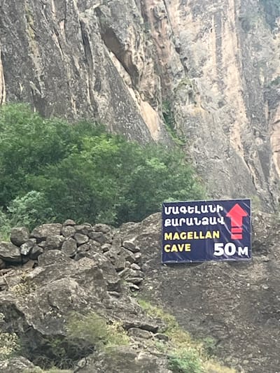 The image depicts a rugged natural landscape in Arpi, Armenia, dominated by a steep, rocky cliff face. The background features a tall, greyish-brown rock formation characterized by prominent vertical fissures and varying textures. A dense growth of vibrant green deciduous trees and bushes is situated at the base and mid-sections of the cliff, primarily on the left and central parts of the frame. In the lower left quadrant, an accumulation of dark grey, irregularly shaped rocks forms a low, rough wall or embankment. A rectangular dark blue sign is affixed to a large, textured rock in the lower right. The sign displays "ՄԱԳԵԼԼԱՆԻ ՔԱՐԱՆՁԱՎ" and "MAGELLAN CAVE" in yellow capital letters, accompanied by a large red upward-pointing arrow and the distance "50M" in white text, indicating the direction and proximity of the Magellan Cave. No people or animals are visible within the scene.