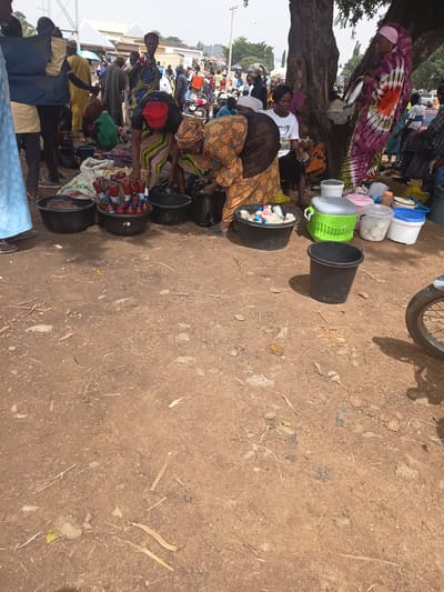 This image captures a bustling market scene in Tilde, Nigeria. A variety of people are present, engaged in selling and browsing goods. In the foreground, several large black plastic basins are arranged on the dirt ground. One basin contains a collection of what appear to be peanuts or nuts. Another basin holds multiple red-bottled condiments, likely cooking sauces or spices. A third basin contains various items, including what looks like bottled water and other packaged goods. Next to these, a bright green plastic cooler is visible, as well as several white and blue plastic containers. A dark gray plastic bucket sits empty to the right of the cooler.

Behind these foreground items, several individuals are interacting. A woman wearing a red headscarf and a striped green and black garment is bent over, attending to goods in one of the basins. Another woman, dressed in an orange and brown patterned outfit, is also engaged with the items in the basins. Further back, a woman in a vibrant tie-dye dress with pink and green hues is standing near a large tree, holding a silver tray. Other market-goers and shoppers can be seen in the background, along with stalls and structures suggesting a lively commercial area. The ground is a mixture of packed dirt and scattered debris, indicating an outdoor marketplace. The overall impression is one of everyday commerce and community activity within the city.