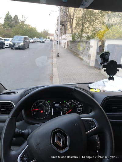 The image is taken from the interior of a Renault vehicle, likely a van, parked on a street in Okres Mladá Boleslav, Czechia, on April 14, 2026, at 07:42 AM. The steering wheel features the Renault logo prominently. The dashboard instruments show a speedometer, a tachometer, and a digital display indicating a temperature of 10°C and an odometer reading of 61812 km, with a trip meter reading of 3367.8 km. A cell phone holder is mounted on the windshield, obscuring some of the view. A car document and a yellow canister are visible.  Outside, a row of parked cars lines the left side of the street, including a Mercedes-Benz van. Buildings and trees are visible in the background. The road appears wet, indicating recent rain. The sky is overcast, suggesting a cloudy morning.