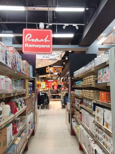 This media file captures a wide-angle, eye-level view of a brightly lit retail store aisle, specifically a home goods or kitchenware section, located in Sindanglaya, Indonesia. The perspective looks down the length of the aisle, with shelves filled with products on both the left and right sides.
On the left, shelves display various kitchen appliances and packaged goods. Items like rice cookers, blenders, and other boxed products are visible, with brand names like "MASPION" and "CROWN" discernible on some packaging. The shelves appear to be a mix of metal and wooden constructions.
On the right, similar multi-tiered shelves are stocked with glassware, including clear jars, drinking glasses, and other packaged household items. The products are neatly arranged, suggesting a well-maintained store environment.
Overhead, a red square sign with white text prominently hangs, reading "Rumah Ramayana". "Rumah" means "Home" in Indonesian, indicating this is likely the home goods department of the "Ramayana" department store chain. The ceiling is dark, featuring fluorescent tube lights and smaller spot lights illuminating the aisle.
Further down the aisle, the store opens up into other sections. Several signs are visible in the background: one partially obscured sign reads "Selesai Pembayaran di sini" (meaning "Finish payment here" in Indonesian), suggesting a checkout area or payment point. Other yellow and red signs indicate promotions or prices, with text like "+ 10%" and "200.000" (likely referring to a discount or price in Indonesian Rupiah). Apparel displays and other merchandise can be seen deeper within the store. The floor is light-colored and clean.