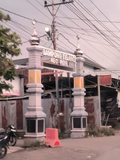 The image displays an entrance gateway on a roadside under an overcast sky, likely marking the entry to a village or district. The gateway consists of two substantial pillars, predominantly grey and white with yellow decorative panels in their mid-sections. Each pillar is topped with a silver, domed finial featuring a crescent moon symbol.

A sign spans between the two pillars, clearly displaying the text "GAMPONG TIBANG" and "KEC. PIDIE". Numerous utility wires are visible overhead, extending from a tall wooden utility pole situated to the right of the gateway. This pole also supports a bright street light.

To the left of the gateway, several motorcycles are parked on the side of the road. On the right, near the base of the right pillar, there is a bright orange/red plastic road barrier. The background reveals buildings with corrugated metal walls and roofs, contributing to a utilitarian aesthetic. Patches of green vegetation grow around the base of the pillars and along the roadside. The scene appears to be daytime, with no people visible, suggesting a quiet moment in this locale within Tibang, Indonesia.