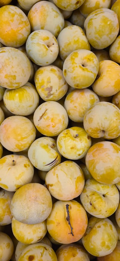 A close-up, top-down view displays a large pile of ripe, yellow plums. The fruits are round to oval-shaped, with a smooth skin exhibiting variations in color, ranging from pale yellow to a more intense golden hue, interspersed with lighter, almost white patches and subtle reddish-brown blemishes. Some plums show a faint line indicating where they would split to reveal the pit. A few have visible stems, and one in the lower right has a small, dark, twig-like stem attached. The plums are packed tightly together, creating a textured, overflowing mound. This scene is a generic market display and does not contain identifiable landmarks or elements specific to Navalcarnero, Spain.
