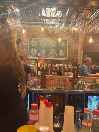 The image depicts the interior of a bustling bar or restaurant in Marquette-lez-Lille, France, characterized by an industrial-rustic aesthetic. The scene is dominated by a long, stainless steel bar counter extending from left to right, laden with various beverage preparation items. In the immediate foreground, bottles with clear, pink, and dark liquids are visible, along with multiple white plastic dispenser bottles topped with red pourers, and a yellow bottle cap.
Behind these items, centered on the bar, is a row of at least nine beer taps, each featuring a brown wooden column and a metallic faucet, implying active beverage service. Clear plastic cups and stacks of orange-tinted cups are arranged near the taps. To the right, an ice bucket with tongs and fresh green herbs (likely mint) is present, alongside bottles of spirits and water.
Several individuals are present. In the extreme left foreground, the back of a person with long brown hair, wearing a dark short-sleeved shirt, is visible. Behind the bar, a man with glasses and short hair is operating the taps. Further right, a man with gray hair, wearing a blue collared shirt, leans against the bar, holding a drink with ice. Additional blurred figures, likely patrons, are visible in the background on the right.
The background features an exposed brick wall adorned with a large, dark chalkboard. The chalkboard displays illustrated circular sections, each containing text in French and numbers (e.g., #1, #2, #3), indicating a menu or selection of beers. Overhead, exposed Edison-style light bulbs hang at varying heights, illuminating the space with a warm glow. A large, ornate chandelier with multiple light sources is suspended from the ceiling. Additionally, a metal rack above the bar holds numerous inverted wine glasses and other glassware. A prominent wooden support beam is visible on the right side of the frame, further contributing to the rustic ambiance.