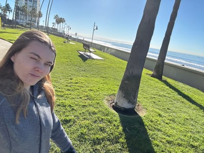 A sideways shot depicts a woman in a grey jacket. She faces the camera with one eye closed. Behind her, a lush green lawn stretches towards a beach and the ocean. Palm trees line the horizon. The sunlight casts shadows from the trees onto the grass. Benches are scattered across the lawn. Buildings rise up in the background, blending with the palm trees.