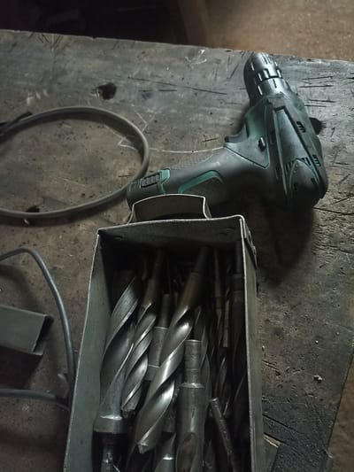 A cordless drill with a green accent is resting on a rough, wooden surface. Next to it, a metal box holds a collection of various drill bits. The wooden surface is worn and appears to be a workbench. The scene is dimly lit, suggesting an indoor or shaded outdoor environment. No people are visible in the image, and no specific location in Polatsk, Belarus, can be determined from the visual information provided.