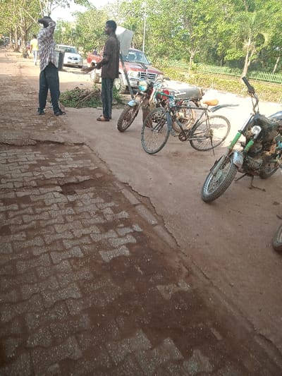 The image shows a paved road in a residential area in Otta, Nigeria. The road surface is a mixture of asphalt and a faded, exposed cobblestone pattern. There is a noticeable crack dividing the road in two sections.  Two men are standing on the left side of the road.  One man is wearing a checkered shirt, a pair of blue jeans and is holding a bottle of water. The other man is wearing a dark t-shirt, a pair of khaki pants, and is holding a broom. A red pickup truck is parked on the right side of the road and two motorcycles are parked on the right side of the road, facing each other. The motorcycles are both in good condition. The image has a sunny outdoor setting with lush greenery, a few trees and some houses in the background. There is an electric wire pole located on the right of the road in the background.  The location of the image is approximately 6.682° N, 3.168° E.